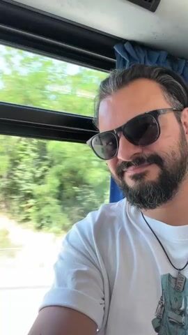 A man with a beard and sunglasses is taking a selfie from inside a vehicle. He is wearing a white t-shirt and a necklace. Outside the window, trees and a road are visible. The vehicle appears to be in motion. The man smiles at the camera.