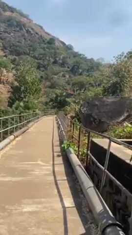 The video shows a man standing on a concrete bridge with metal railings. He is facing a hillside covered in sparse vegetation and large rocks. The sky is blue with some clouds. The man is wearing a light purple and white checkered shirt and sunglasses. The bridge appears to be part of a dam or water management system.