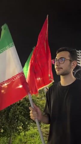 A young man wearing glasses and a black t-shirt stands holding two flags on poles at night. The flag in the foreground on the left is predominantly white and red, with green at the top. The flag behind it on the right is red with some emblem or writing. The background is dark, suggesting nighttime, with some greenery visible to the left and a light fixture or structure behind the flags. The man is looking off to the right with a serious expression. The flags are waving slightly.