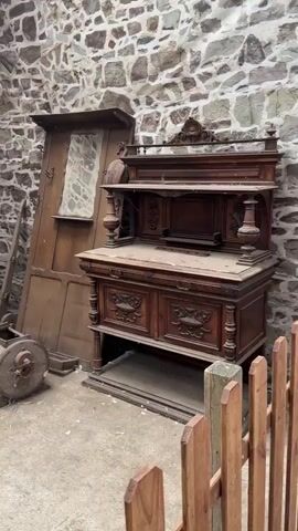 The video begins by showing a rustic stone-walled room with various antique items. To the left, a wooden ladder leans against a large, upright wooden door that has a reflective surface, possibly a mirror. Beside the ladder is a round, wooden barrel with metal accents. In the center of the room is a large, ornately carved wooden cabinet, likely an antique dresser or sideboard. Further into the room, a vintage piano with a wooden bench in front of it is visible. A large, brown teddy bear is seated on the bench, facing the piano. To the left of the piano, a metal and wood cart with yellow wheels and a red seat is present. A light-colored jug sits on the ground near the cart. The camera then pans to the right, revealing a wooden fence in the foreground. The scene shifts to a larger barn-like structure with a wooden ceiling and stone walls. In this space, several wooden benches and small wooden fences are arranged. A person, wearing a hat and a brown shirt, stands on the right side of the frame, facing a yellow table with a game laid out on it, possibly a shuffleboard or a similar table game with small brown objects. The person's hands are extended as if playing or demonstrating the game. The camera continues to pan, showing more of the interior of the barn and the games laid out on tables. The floor of the barn appears to be dirt or gravel.