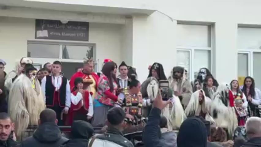 A group of people, some in costumes, are standing on steps of a building with a sign written in Cyrillic. The costumes include a king, a person in colorful clothing, and others in white, furry garments and masks.  People in the crowd are taking pictures with their phones.  The sign on the building says "НАРОДНО ЧИТАЛИЩЕ Отец Паисий 1927".
