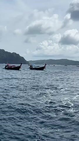 A medium shot captures a body of water with several boats dotted across the surface. The water is a deep blue and appears choppy with white foam. In the foreground, the wake of a boat creates a foamy trail. In the middle ground, two long, narrow boats with pointed ends and raised prows are anchored. These boats have wooden structures on them. In the background, more boats are visible, some appearing smaller and further away. The horizon is lined with a hazy green landmass, likely an island or coastline, covered in lush vegetation. The sky above is a mix of grey and white clouds, suggesting an overcast or partly cloudy day. The overall scene depicts a serene coastal environment with typical Thai longtail boats.