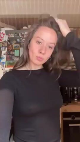 A woman in a black long-sleeved shirt and light-colored pants is standing in a kitchen. She is facing the camera and messing with her hair. She puckers her lips, then folds her arms across her chest, turns to the side, and looks at the camera again. The kitchen has light-colored cabinets and a refrigerator covered with magnets and pictures. There are dishes in the cabinets. An oven is also visible.