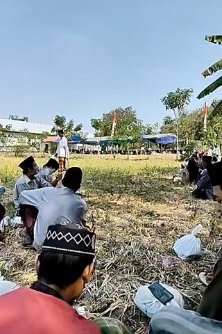 A group of men are gathered outdoors, some sitting and some standing. One man in a white long-sleeved shirt and a patterned sarong stands in the middle distance. Several men in the foreground are wearing black skullcaps, and one man wears a green skullcap. The setting appears to be a grassy area with some trees and buildings in the background. A flagpole with a flag is visible on the right side of the frame. The men appear to be engaged in a shared activity or observation, though their specific actions are not clearly discernible from the still images.