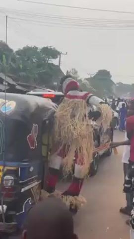 A video depicts a person dressed in a traditional costume and mask walking amidst a crowd of people on a street. The costume has a white and red striped torso and a skirt of straw-like material. People stand around, observing, and one person guides the costumed figure by the arm. There are vehicles parked on the side of the street and overhead power lines are visible. The camera viewpoint is unsteady, and at times the ground is visible. The weather appears to be overcast.