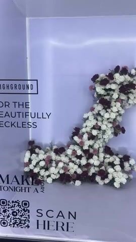 This media file appears to be a vertical video capturing a floral display in what looks like an event setting or photo booth. The primary subject is a large, abstract floral arrangement composed of white and deep red/burgundy roses, with some smaller pink flowers interspersed. The flowers are densely packed, creating a flowing, almost sculpted shape that occupies a significant portion of the frame.

The setting is a clean, modern backdrop with a soft lavender gradient. There is visible text on the backdrop, suggesting it's part of a branded event or promotion. On the left side, large black lettering spells out "HIGHGROUND" at the top. Below this, in a smaller black box, is the word "HIGHGROUND" again. Further down, there are lines of text: "FOR THE", "BEAUTIFULLY", and "RECKLESS". At the bottom left, there's a QR code and the text "MAKE TONIGHT A STORY" and "SCAN HERE".

On the right side of the backdrop, more text is visible in black lettering. At the top, it reads "ROOFTOP KISSES & UNTOLD STORIES". Below this, some text is partially obscured but appears to mention locations or themes, with "PHNOM PENH" and "MOST DANGEROUS VIEW" being legible. Near the bottom right, "SKYBAR" is visible above a barcode-like graphic.

The lighting appears to be artificial and bright, creating a slight glow around the edges of the backdrop. There are no people clearly visible within the frame. The overall mood is celebratory and sophisticated, likely associated with a night-time event. The location context provided is Khan Boeng Keng Kang, Cambodia, which, combined with the text mentioning Phnom Penh, places the event in the capital city of Cambodia. The time of day is likely evening or night, given the artificial lighting and the "night" reference in the text.