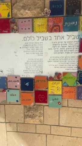 This video shows a wall adorned with numerous colorful, small square tiles, many of which contain writing and drawings. The tiles are arranged in rows and columns, creating a mosaic-like effect. Some tiles feature Hebrew text, while others display simple graphics like hearts, rainbows, and geometric shapes. A larger white board or poster is partially visible in the upper right quadrant of the frame, also featuring text. The wall itself is constructed of beige, rectangular stone blocks. The lighting suggests an outdoor setting, possibly a walkway or courtyard, with shadows cast on the wall. There are no people visible in the video.
