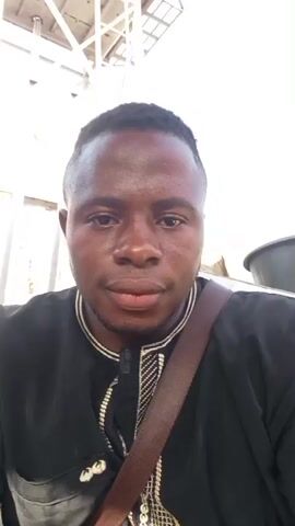 This is a close-up selfie video of a Black man wearing a black robe with intricate gold embroidery around the collar and down the front. He has a short haircut and is looking directly at the camera with a neutral expression, though his eyes appear a little weary. A brown strap from a bag or satchel is visible crossing his chest. The background is slightly blurred but suggests an outdoor setting with some light-colored structures and possibly some greenery at the bottom. The lighting is bright, indicating it is daytime, likely sunny. There is no discernible text visible in the video. The overall impression is of a person taking a casual selfie outdoors in Nigeria.