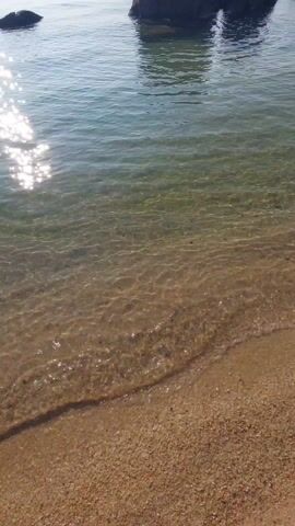 The video captures a serene beach scene with clear, shallow water gently lapping onto a sandy shore. Several large, dark rocks are visible in the water, some close to the beach and others further out. The water is so clear that the sandy bottom is easily visible. The sun glints off the water's surface, creating a sparkling effect. In the background, a distant landmass is visible, along with a ship or boat. The sky is blue with a few scattered clouds. The overall impression is one of tranquility and natural beauty.