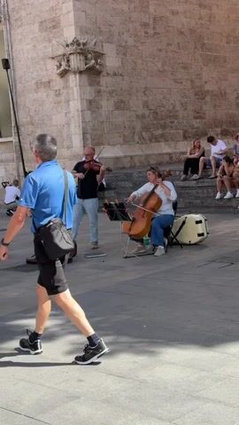 Two musicians are playing string instruments on a paved area in front of a stone building. On the left, a bald man wearing glasses, a black t-shirt, and light blue jeans stands and plays the violin. His violin case is open on the ground to his left. A music stand is positioned in front of him. On the right, a woman with brown hair, wearing a white long-sleeved shirt and blue jeans, sits and plays the cello. A music stand is in front of her. A large white drum is behind her to the right. Several people are seated on stone steps behind the musicians. Some are watching the performance. A bicycle is visible to the left of the musicians, behind the violinist. The sun is shining, casting shadows.