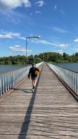 Two individuals walk on a wooden bridge over a body of water. The person in the foreground, facing away from the camera, is wearing a black shirt, shorts, and boots, and is carrying a bright orange object, possibly a ball. Further down the bridge, another individual, facing the camera, is wearing a light-colored shirt and dark pants. The bridge has a white railing on both sides and is lined with lampposts. Lush green trees and vegetation are visible on the distant shoreline under a bright blue sky with scattered white clouds. The camera is positioned behind the first individual, looking down the length of the bridge.