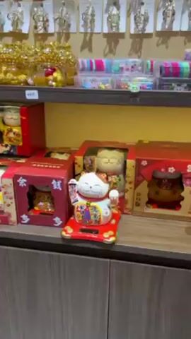 The video shows a shelf in a store displaying several Maneki Neko figurines, also known as lucky cats. The figurines are mostly white with colorful details, including Chinese characters, and are sitting on red platforms. Some of the figurines are still in their boxes, while one is displayed on the shelf. The background is a light yellow wall, and there are other items on the shelf above the figurines, including golden objects in glass jars and colorful hair accessories. The camera zooms in on the displayed figurine. The figurine’s arms are raised and adorned with scrolls. The platform is powered by a solar panel on the front, causing the cat to wave.