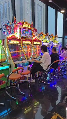 The video shows a row of arcade games in what appears to be an arcade or entertainment center. The games are brightly lit with neon lights and colorful displays. In the foreground, there are two empty orange chairs. Several people are playing the arcade games. The person closest to the camera is a young man wearing a white t-shirt and black pants, sitting and playing one of the games. To his right, another person is playing a game, and further down the row, more people are visible engaged in playing. The arcade games have large screens displaying graphics, and some appear to be racing or carnival-themed games with titles like "CRAZY CLOWN." The environment is characterized by a shiny, reflective floor and large windows in the background showing an urban building. The overall atmosphere is one of entertainment and leisure.