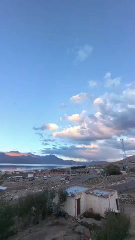 The video shows a wide shot of a village next to a large body of water with mountains in the background. The sky is blue with scattered clouds that are tinged with pink and orange, suggesting it is either sunrise or sunset. In the foreground, there are a few small buildings, some dirt roads, and sparse vegetation. There are also a few cars parked in the village. The overall impression is of a remote and peaceful settlement in a mountainous region.