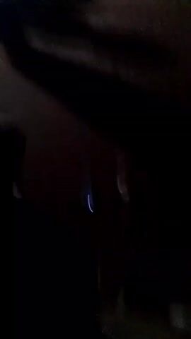 A person is walking toward a dimly lit gathering of people sitting in chairs outdoors. A bright light illuminates the area where people are sitting. The person continues walking past the gathering, along a dirt surface with shadows. The video is shaky.