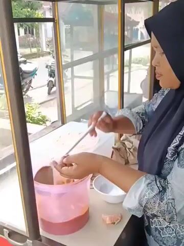 A person wearing a hijab and a patterned shirt is standing behind a food stall with glass windows. They are using a ladle to scoop a reddish-brown liquid from a large pink container into a clear plastic bag. The person is holding the bag with one hand while the ladle is in the other. There is a small white bowl and some brown paper bags on the counter. Outside the stall, a motorcycle and some greenery are visible.