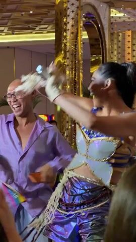 In a vibrant indoor setting, likely a performance venue or event space, two individuals are the center of attention. The person on the left, with a shaved head and glasses, is wearing a loose-fitting lavender button-up shirt and flowing light blue pants. They are actively engaged in a dynamic movement, their arms outstretched and their body angled as if dancing or gesturing enthusiastically.

To their right, a person with long, dark hair pulled back into a high ponytail is dressed in a striking outfit. They wear a beige and blue patterned top with a corset-like design, cinched at the waist. Below, a voluminous skirt or fabric drapes, shimmering with iridescent purple and blue hues. Their arms are also in motion, with one hand holding what appears to be a microphone or prop, and the other adorned with feathery accessories. This person is leaning forward, their expression conveying energy and perhaps a musical performance.

Several spectators are visible in the foreground, seated and observing the performance. Their faces show a range of engagement, from attentive to smiling. The background reveals a richly decorated environment with polished floors, ornate golden architectural elements, and some greenery. The lighting suggests an indoor event, possibly during the evening or under artificial illumination. Slot machines are visible in the distance, hinting at a casino or entertainment complex setting within Cebu City, Philippines. The overall atmosphere is lively and celebratory, suggesting a performance or show in progress. No visible text is present in the frames.
