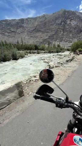 A first-person perspective video shows a motorcycle rider on a paved road next to a riverbed. The rider is wearing a black jacket, black gloves, and a yellow and burgundy helmet. They are holding onto the handlebars, and the camera, mounted on the motorcycle, is pointed forward. To the left of the road, a wide, rocky riverbed is visible, with a fast-flowing river running through it. Beyond the riverbed, there are trees and steep, rocky mountains under a blue sky with scattered clouds. The motorcycle has a red and white fuel tank and a circular mirror reflecting the rider holding up a smartphone. The road appears to be in a remote, mountainous region.