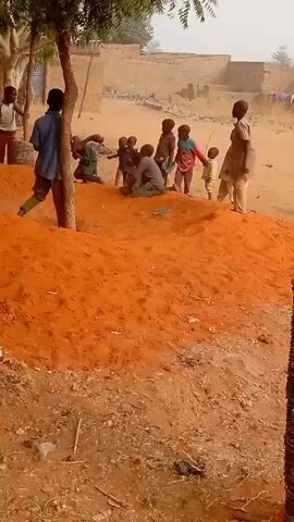 Several children are gathered around a mound of reddish-orange soil in an outdoor setting. Most of the children appear to be standing or sitting on the mound, while at least one is playing near the base. A man in a blue shirt and green pants is near a tree, apparently interacting with the children. There are scattered trees, sparse vegetation, and dry dirt ground around the mound. In the background, simple buildings are visible, and a goat is seen grazing nearby. The atmosphere looks dry and possibly dusty, with a somewhat hazy sky.