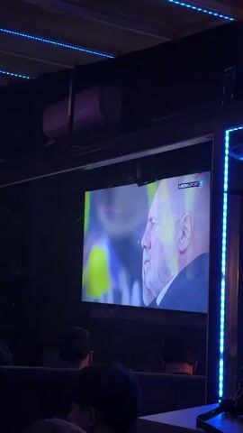 The video shows a group of people gathered in a dimly lit room, likely a bar or lounge, with neon lights lining the ceiling. A large television screen displays a man in a suit and what appears to be a sports broadcast. Several individuals are seated, with some standing and moving around. The atmosphere seems relaxed, with people engaged in conversation and watching the screen. The camera pans across the room, capturing different angles of the gathering and the environment. There are drinks and personal belongings visible on tables.