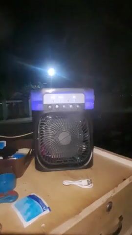 A black portable air cooler with blue lighting is sitting on a wooden surface. The cooler has a fan in the center and buttons on top. There is a white USB cable on the surface in front of the cooler, along with two blue ice packs. The background is dark, suggesting it is nighttime, with a bright light source visible in the distance. The air cooler is emitting mist.