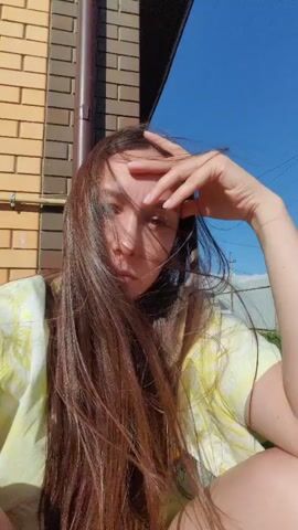 A woman with long, dark brown hair sits outside, her hair blowing in the wind. She wears a yellow and white tie-dye shirt. Her right hand is raised to her forehead, and her eyes are partially obscured by her hair. Behind her, a brick wall and a drainpipe are visible. The sky is bright blue.