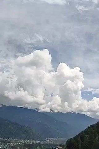 The video captures a scenic view of mountains and a cloudy sky. The foreground shows a dense green forest, with a small town or village nestled at the base of the mountains. The mountains themselves are covered in lush vegetation, with some areas appearing darker, possibly indicating deeper valleys or shadows. Above the mountains, a dramatic sky is visible, filled with large, fluffy white clouds that are illuminated by sunlight. Some thinner, wispy clouds stretch across the upper portion of the frame, creating a textured effect against the blue sky. The overall impression is one of a serene and natural landscape, with the clouds adding a dynamic element to the scene.