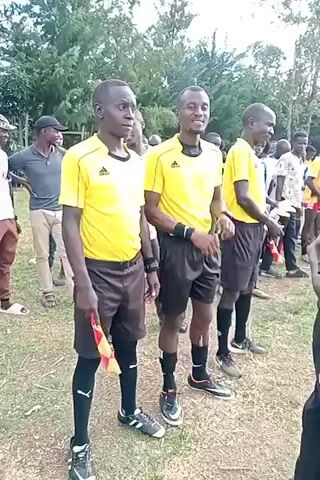 Three men in yellow referee shirts and dark shorts are standing side-by-side in a grassy field. The man in the center smiles slightly. They are wearing black socks and sneakers. The man on the left is holding a red and yellow flag. Behind them are other people, some in sports uniforms. A man in a dark grey shirt and hat is talking to a man in a white shirt, who is holding a piece of paper. The environment appears to be a park or field with trees in the background.