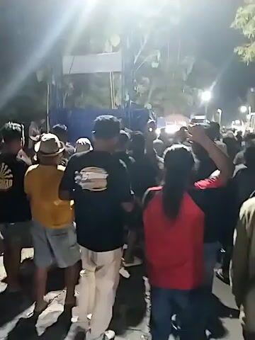 This video captures a nighttime outdoor gathering in Krebet, Indonesia. A crowd of people is visible, many of them wearing casual clothing. Some individuals are wearing hats, including a light-colored fedora-style hat on a person in a yellow shirt. The lighting suggests it is nighttime, with bright artificial lights casting a glow on the scene and creating lens flares. In the background, there appear to be tents and possibly vendor stalls, indicating some kind of event or market. Trees are visible, suggesting an outdoor, possibly tropical, setting. The overall mood seems to be energetic, with people standing, looking around, and some with their arms raised, possibly dancing or engaging with the event. There's a sense of movement and a lively atmosphere. Some visible text on a person's shirt in the foreground appears to be a logo or graphic with possibly some lettering.