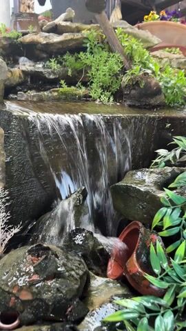 The video showcases a small, man-made waterfall feature within a garden setting. Water cascades down a rocky facade, creating a miniature waterfall effect. Plants are strategically placed around the waterfall, adding to the natural aesthetic. A small clay pot is positioned near the base of the waterfall, partially submerged in the water. A wooden barrel planter containing purple flowers sits in the foreground, slightly out of focus. In the background, steps can be seen leading up to a raised platform or deck, further enhancing the garden environment. The overall scene evokes a sense of tranquility and natural beauty.