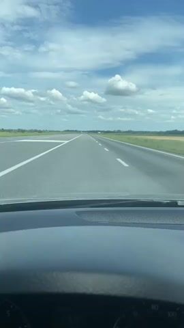 This is a first-person perspective video taken from inside a vehicle driving down a long, straight road. The road appears to be an airport runway, indicated by the wide expanse of asphalt and the white lines marking lanes. The scene is set in a rural area, with flat, green fields visible on either side of the road, extending to the horizon.

The sky is a vibrant blue, dotted with fluffy white clouds, suggesting a fair weather day, likely daytime. The sun is shining, casting a bright light on the scene. The vehicle's dashboard is partially visible at the bottom of the frame, showing some instruments, including a speedometer that indicates the vehicle is moving. There is no visible text in the scene other than the markings on the road and the dashboard.

The overall mood is one of travel and openness. The long, clear road and expansive sky create a sense of freedom and journey. The lack of other vehicles or people suggests a quiet and possibly secluded environment. The setting in Balivka, Ukraine, adds a specific geographical context to this open, likely rural landscape.