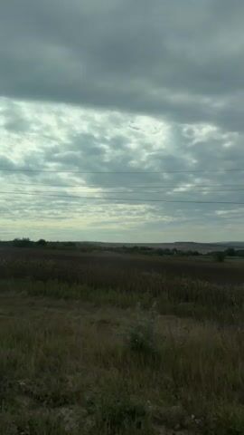 A drone or camera on a vehicle moves across a rural landscape under a cloudy sky. The terrain is mostly flat with fields of dry grass and crops. There are some trees and a power line pole visible. The sky is overcast with grey clouds. The drone/camera appears to be moving from left to right.