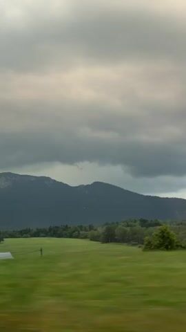The video captures a blurred view of a rural landscape from a moving vehicle. In the foreground, green foliage and tree branches streak across the screen, indicating motion. Behind them, a wide, flat field of vibrant green grass stretches towards a line of trees. Several buildings with red-tiled roofs are visible in the middle ground. In the background, rolling hills rise, culminating in a darker, more mountainous range under a cloudy sky. The clouds are heavy and gray, suggesting an overcast or potentially stormy day. The overall impression is of a quick journey through the countryside.