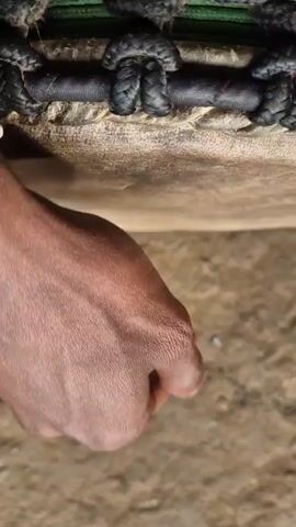 The media file is a close-up, vertically oriented video showing a person's hand playing a drum. The drum appears to be made of natural materials, possibly animal hide, with a rough, textured surface. It is reinforced with a woven black cord along the top edge. The hand, which is dark-skinned, is shown striking the drum with a closed fist or the knuckles. The background is a sandy or earthy surface, suggesting an outdoor setting. The lighting appears to be natural, possibly daylight, but there are no clear indicators of the time of day or weather conditions. The overall mood is focused on the action of drumming, with the sound of the strikes being the primary sensory input. The context provided states the location is Shila, India. There is no visible text in the media file.