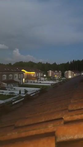 The video shows an aerial view of a residential area as dusk settles. The foreground is dominated by a close-up shot of a terracotta tiled roof. In the mid-ground, a row of multi-story townhouses with brown roofs and light-colored facades are visible. Many of the windows are illuminated, suggesting people are home. Some townhouses have outdoor lighting, including string lights above one of the entrances. Greenery, including trees and bushes, surrounds the houses and forms a treeline in the background. The sky transitions from dark grey clouds at the top to a clear blue with streaks of orange and yellow near the horizon, indicating the sunset. The camera appears to be slightly tilted and moves very subtly throughout the clip, creating a gentle panning effect. No people are clearly identifiable in the video.