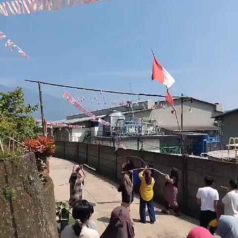 The video shows a group of people, mostly adults, participating in a game on a paved road on a sunny day. There are decorations, including an Indonesian flag and red and white streamers, hung above the road. Some people are holding a long rope, while others are jumping over it. The road is lined with a concrete wall on one side and a slope with plants on the other. In the background, there is a large industrial building with tanks and pipes, along with a mountain in the distance.