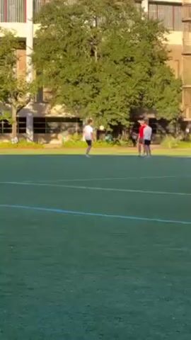 The video shows three young men on a green artificial turf field. One of the men, wearing a white shirt and black shorts, kicks a soccer ball in the direction of the other two men. One of the men is wearing a red shirt. There is a multi-story building and trees in the background. It is daytime and the lighting is bright.