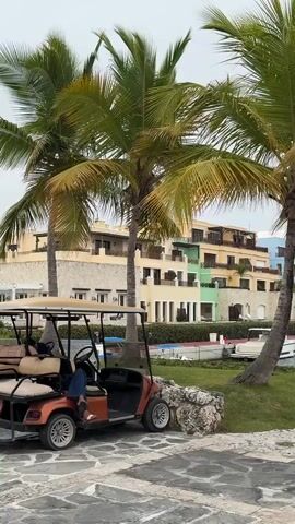 The video shows a golf cart parked on a stone walkway with people sitting inside. Behind the golf cart are palm trees, a grassy area, and a colorful building complex. The building appears to be a resort or hotel, with multiple levels and balconies. Several boats are docked in the water in front of the building. The sky is overcast, suggesting a cloudy day. The overall scene depicts a tropical or coastal vacation setting.