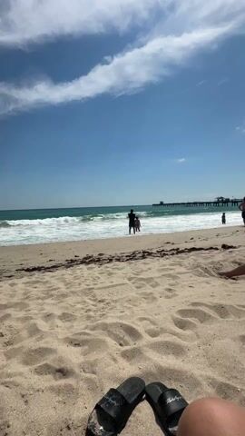 The video shows a beach scene on a sunny day. In the foreground, there's sandy beach with visible footprints. Two black sandals are placed near the bottom edge of the frame. The midground shows a group of people standing at the shoreline, where waves are breaking. A pier stretches out into the ocean in the distance. The sky is blue with scattered clouds.
