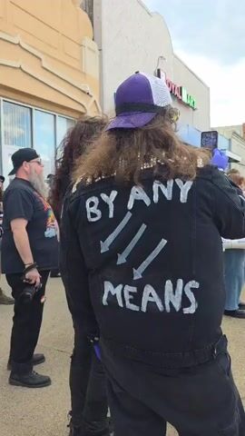 The video shows a small crowd of people standing on a sidewalk in a city. The person in the foreground is wearing a black jacket with the words "BY ANY MEANS" painted on the back along with three downward-pointing arrows, and they are holding a cell phone and taking pictures. Other people are nearby, including a man with a beard and a camera, a person with long curly hair, and other individuals in the background. The crowd is standing in front of a building with a "Royal Mart" sign. The sky is partly cloudy. The scene appears to be a demonstration or protest of some kind.