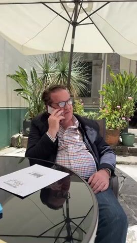 A medium shot shows a man in his 50s talking on a cell phone while sitting at a table outside. The man has fair skin, light brown hair, and is wearing glasses. He wears a multicolored plaid shirt, a dark jacket, and blue jeans. The phone is held to his left ear. He appears to be listening intently to the person on the other end of the call. On the black glass table in front of him, there is a document. Behind him, potted plants and a sun umbrella are visible.