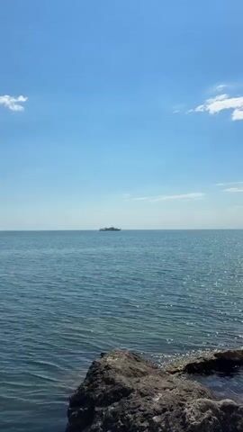 A ship is sailing across the ocean. The sky is blue with a few white clouds. The water is calm and reflects the sunlight. There are rocks in the foreground.