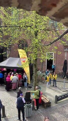 The video shows an outdoor public event in a brick-paved square, crowded with people. A large brick building with a gothic arched entrance stands in the background. A tree with vibrant green foliage stands in the center of the square, partially obscuring the view. A white tent with a black covering houses what appears to be a vendor booth with colorful items. A yellow flag with red lettering and a heart symbol is prominently displayed next to the tent. People are gathered around the tent, some appearing to browse the items on display. Additional people are standing in small groups or walking through the square. A temporary stage is set up next to the church. The footage is shot from an elevated position, looking down into the square, with a portion of a stone structure visible at the top of the frame.