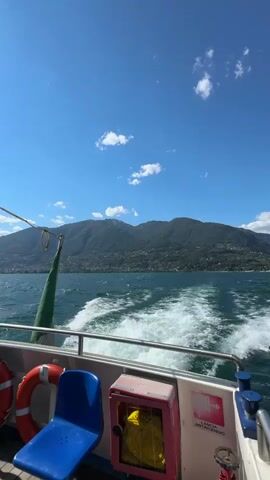 The video shows a view from the back of a boat on a sunny day. The boat is moving, leaving a wake behind it. In the distance, there are green, tree-covered mountains. The sky is bright blue with a few white clouds. On the deck of the boat, there are two blue seats, two orange life preservers, and a red fire extinguisher box. A green flag is hanging from a pole on the boat. The water appears choppy.