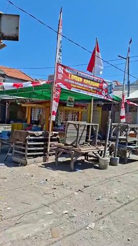 The video pans across a street scene under a clear blue sky. There are several small shops or stalls lining the street, some with awnings. Indonesian flags are displayed, indicating a celebration or national event. Objects visible include wooden benches, carts, motorcycles, a pickup truck with the word "MITSUBISHI" on the back, and various items for sale displayed outside the shops. There are a few people visible, sitting or standing near the shops and on benches. The ground appears to be unpaved, with some debris scattered around. The overall impression is of a local marketplace or street with small businesses operating.