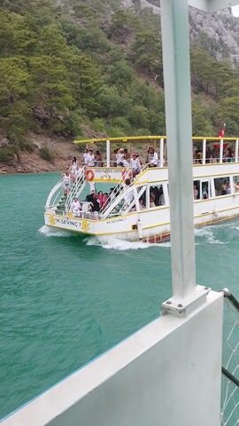This media file shows a wide-angle, high-angle shot of a passenger boat moving through turquoise water. The boat is white with yellow trim and has multiple levels filled with passengers. The passengers appear to be tourists, and many are looking out at the scenery or at the camera. Some are standing on the open deck, while others are seated inside.

The boat is traveling alongside a lush green, forested mountainside, suggesting it is in a canyon or a coastal area with steep terrain. The water is choppy, indicating movement and perhaps a slight breeze. The overall impression is of a scenic boat tour.

The boat is labeled "M. SEVINC 1" and also has text that reads "GREEN CANYON" and "GREEN LAKE" on the side, providing a strong clue to the location. The presence of a Turkish flag on the boat further confirms the location in Turkey. The time of day appears to be daytime, with bright, natural lighting, and the weather seems pleasant for a boat trip. The mood is relaxed and enjoyable, typical of a tourist excursion. The water's vibrant color and the dense greenery of the mountains contribute to a picturesque and inviting scene.