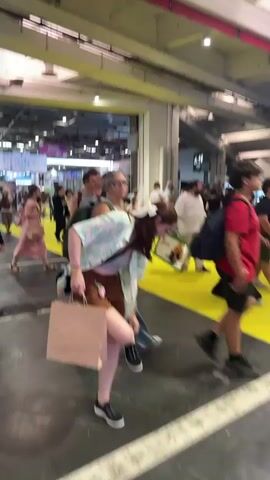 The video shows a bustling indoor event, likely a convention or expo, with numerous people walking and interacting. In the foreground, a woman with long brown hair tied back with a white bow is bending over, holding a brown paper bag. She is wearing a light-colored short-sleeved shirt with a floral pattern, brown shorts, and black sneakers. To her left, two individuals walk past, one wearing a furry animal mask and the other a black dress and a fox-like mask with ears. Further in the background, a crowd of people are seated at tables, some eating and drinking. The environment features bright overhead lighting, concrete flooring, and large open doorways leading to other areas of the venue. A distinctive bright yellow stripe runs along the floor in some sections. The overall atmosphere appears lively and crowded.