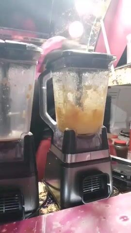 The video shows two blenders sitting on a pink countertop with a speckled pattern. The blender on the left has a mixture of a brownish color inside, while the blender on the right has a lighter, pinker mixture. A woman wearing a black shirt and red pants approaches the blenders. She picks up the blender on the right. The setting appears to be outdoors, possibly at a food stall, with the background suggesting nighttime. String lights are visible in the background. A credit card reader is also visible on the counter.