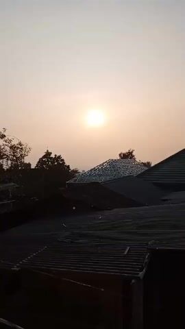 The video shows a view of rooftops and trees against a hazy sky with the sun visible as a bright, diffused orb. The sun is in the upper center of the frame, casting a soft, warm light. The rooftops are dark and appear to be made of corrugated material. Some of the roofs have visible construction elements, such as metal framing, suggesting ongoing work or renovation. Trees with dark foliage are silhouetted against the sky on the left side of the frame. The overall atmosphere is calm and serene, possibly indicating early morning or late afternoon.