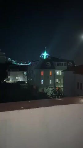 The video shows a nighttime cityscape with buildings and a bright, illuminated structure on top of one of the buildings. The illuminated structure appears to be a Ferris wheel or a similar amusement ride with brightly colored lights, including blue and green. The building below has several windows, some of which are lit with orange and white light, suggesting occupancy. There is a dark, possibly concrete, ledge or railing in the foreground, and the sky is dark with a hint of moonlight visible. The overall scene conveys an urban nighttime environment, possibly a town or city with some form of entertainment or decoration.